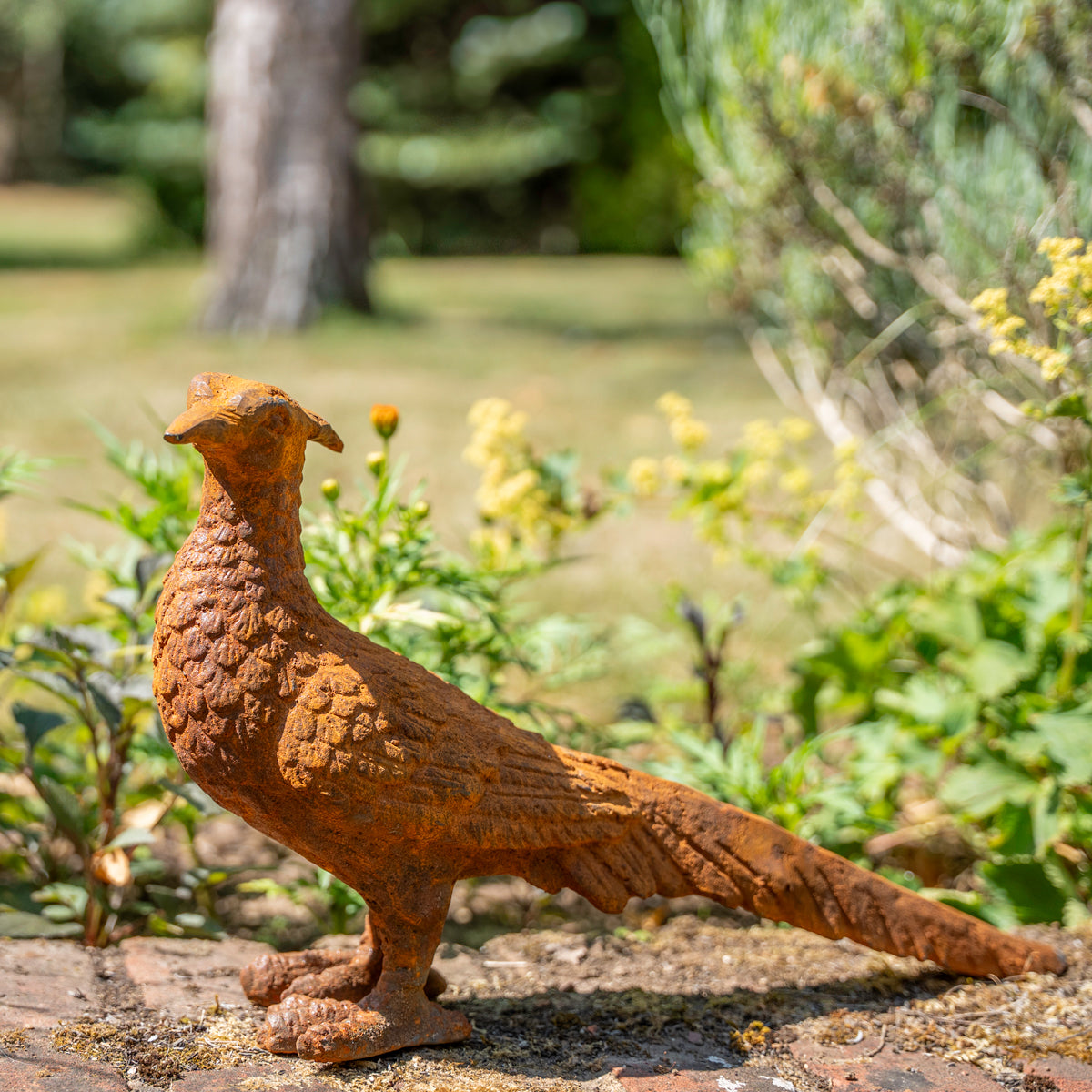 A rusted metal pheasant garden statue stands on a stone surface outdoors, with blurred greenery and yellow flowers in the background.