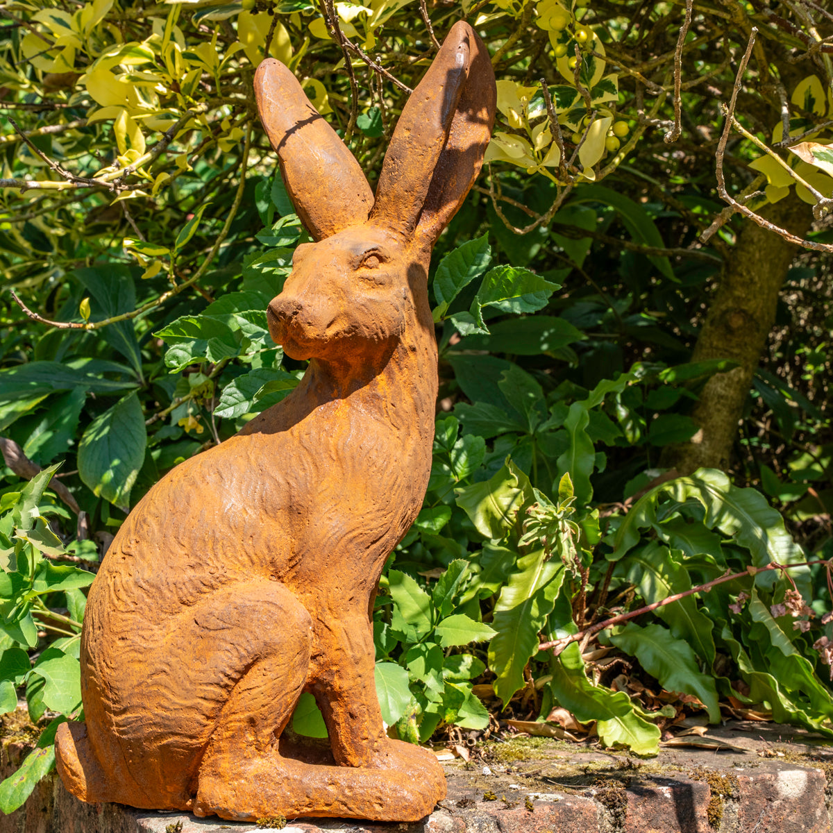 A terracotta coloured iron garden statue of a sitting hare, shown outdoors against a background of green foliage and trees.