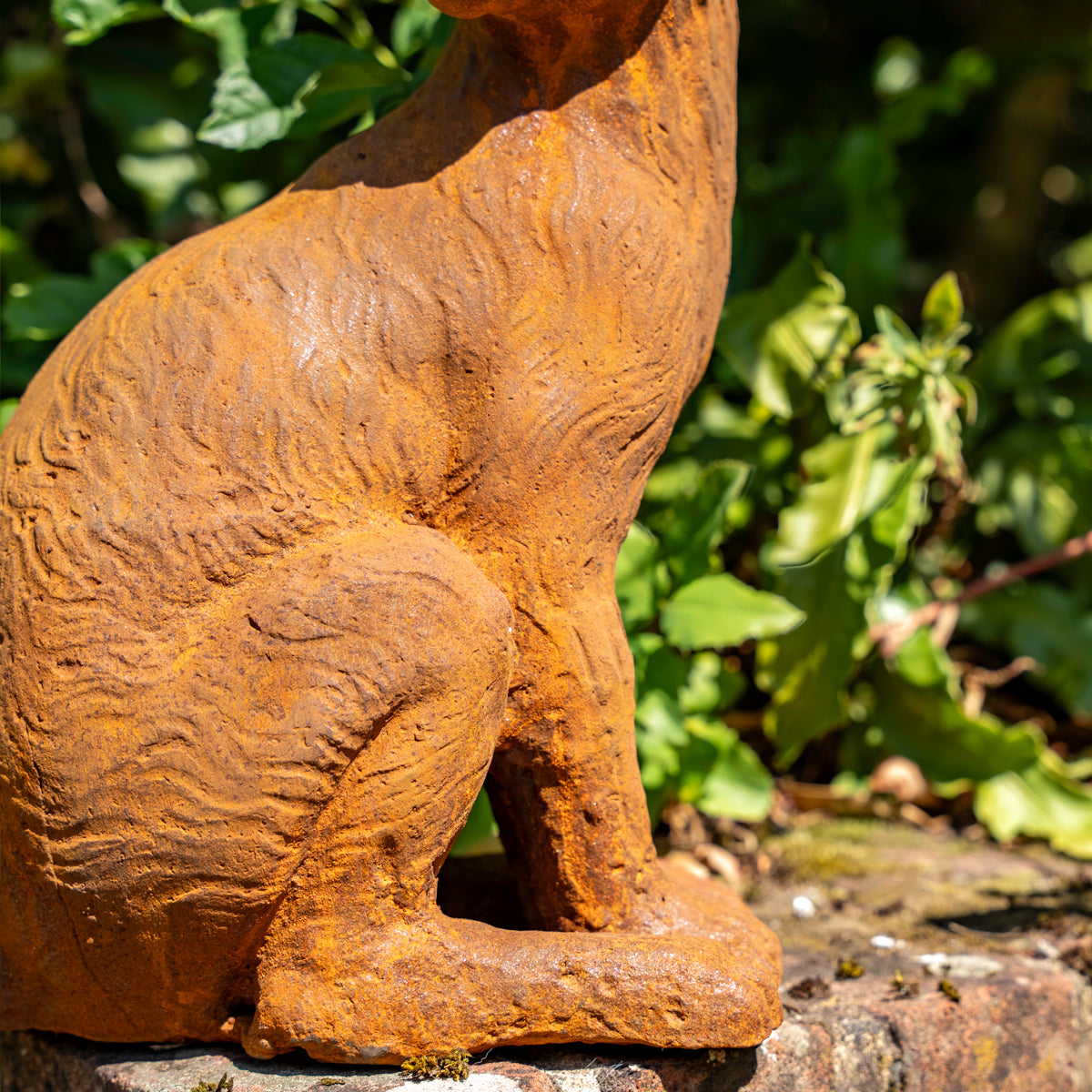A rusted orange-brown metal garden statue of a seated cat, shown outdoors against a blurred green garden background.