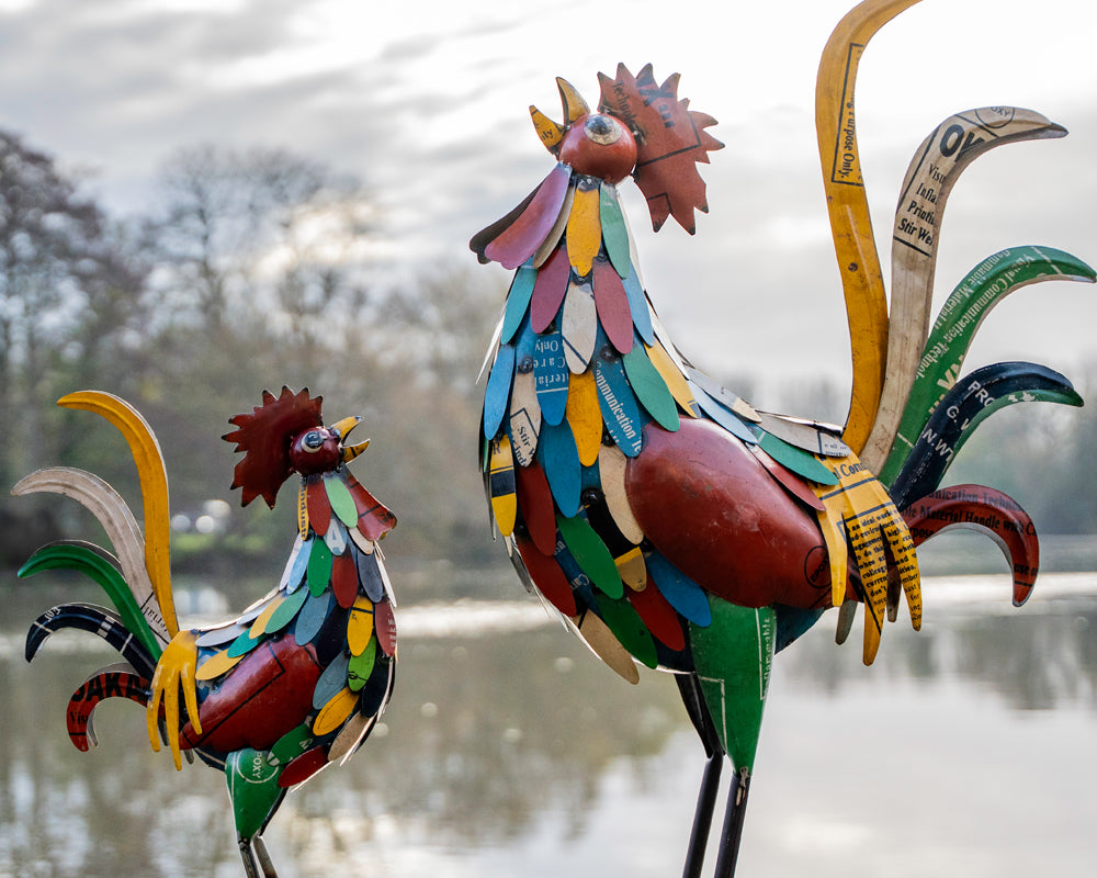 Colorful metal rooster sculptures on a wooden surface with a blurred natural background