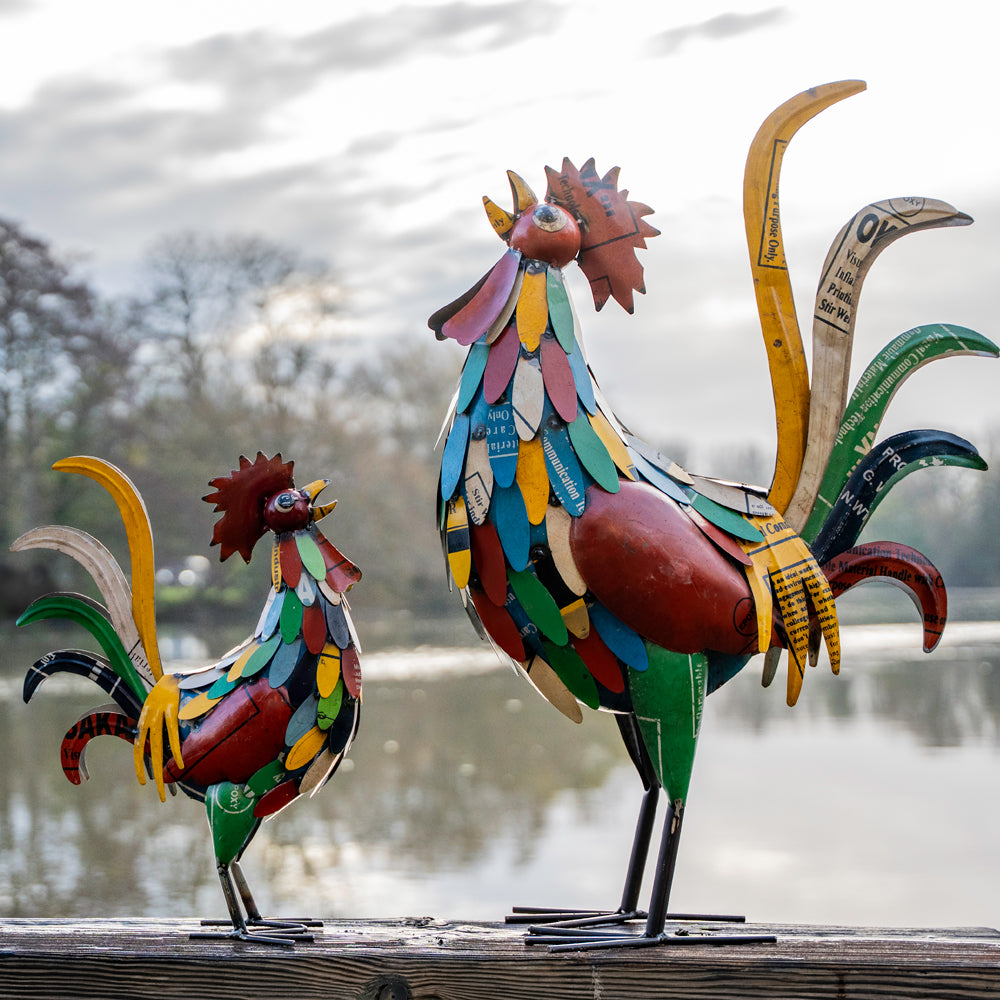 Colorful metal rooster sculptures on a wooden surface with a blurred natural background