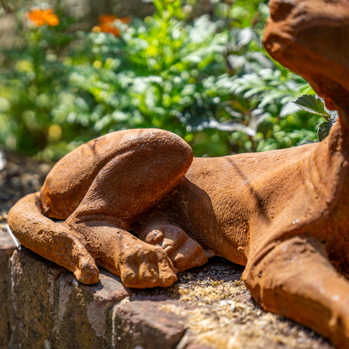 A Rusty Dog garden statue lying down, positioned on a stone ledge with a blurred green garden background.