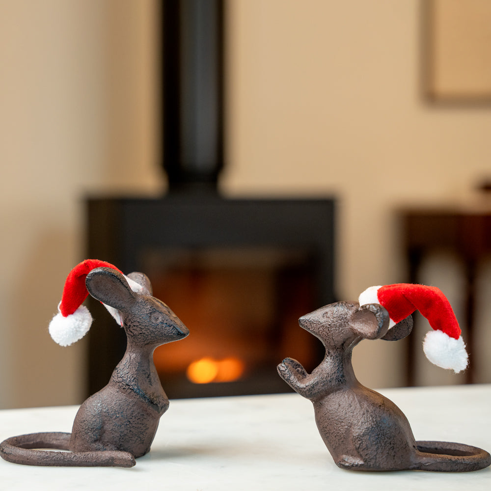 Two cast iron mouse figurines wearing Santa hats in front of a fireplace.