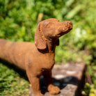 A rustic, rusted metal dachshund statue standing outdoors on a stone slab with a blurred green garden background.