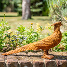 A rusted orange-brown metal pheasant garden statue standing on a brick ledge with a blurred garden background.