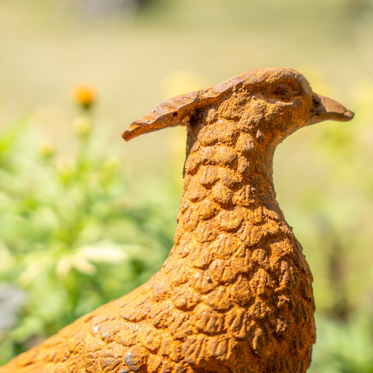 A close-up of a textured bird sculpture or garden statue in a warm rust-orange finish, shown outdoors against a blurred green and yellow background.