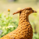 A close-up of a textured bird sculpture or garden statue in a warm rust-orange finish, shown outdoors against a blurred green and yellow background.