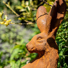 A close-up of a rusted metal hare garden statue with a textured, weathered finish, set against a blurred green garden background.