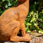A rusted orange-brown metal garden statue of a seated cat, shown outdoors against a blurred green garden background.
