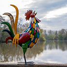 Colorful rooster sculpture on a wooden platform with a lake and trees in the background