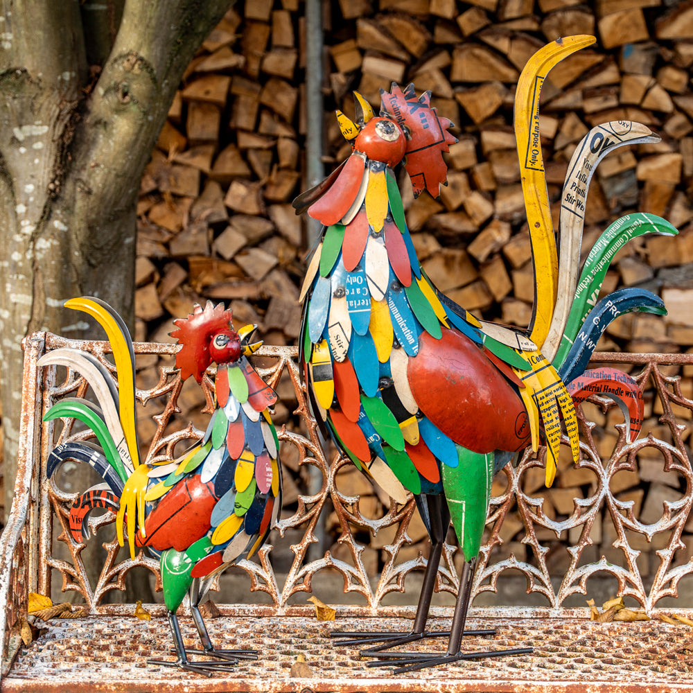 Colorful metal rooster sculptures on a mosaic surface with stacked firewood in the background