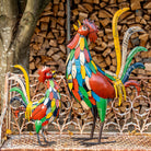 Colorful metal rooster sculptures on a mosaic surface with stacked firewood in the background
