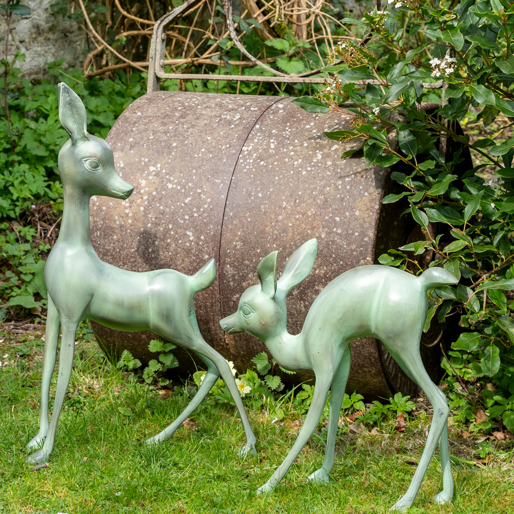 Two metallic deer sculptures in a garden setting with a stone barrel and greenery.