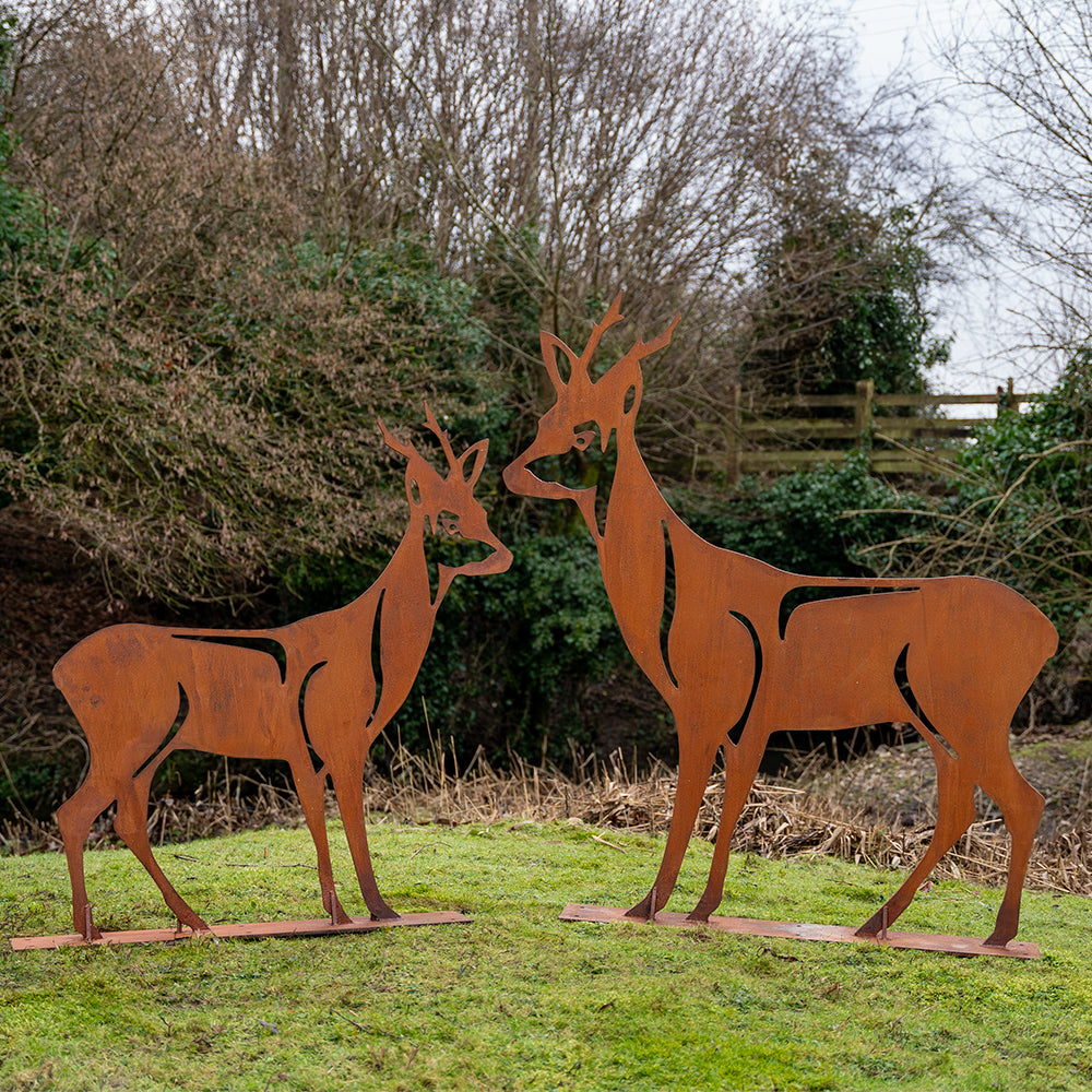 Two brown metal deer silhouettes standing on a grassy field, with trees and a fence in the background.