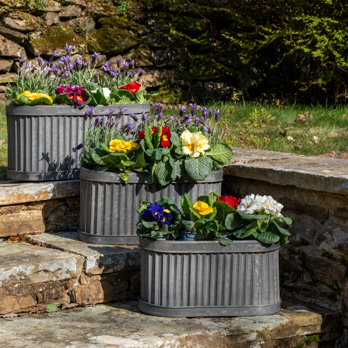 Three gray metal planters with colorful flowers on a stone ledge.