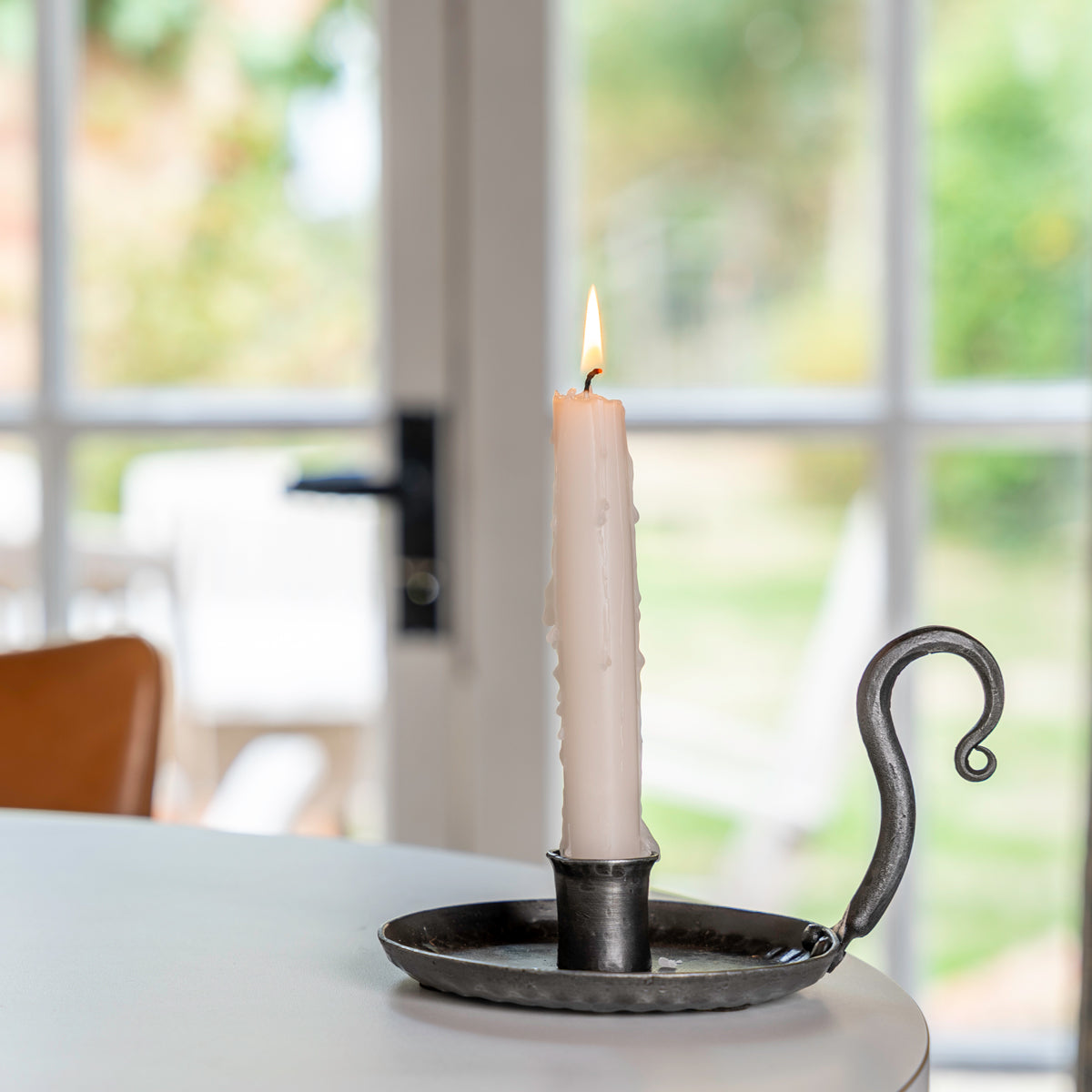A lit white taper candle in a dark metal holder with a curved handle, set on a white table in front of a softly blurred window background.