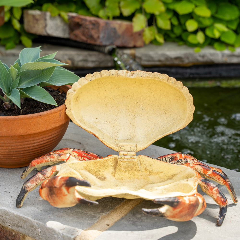 Decorative crab with an open shell on a stone surface near plants and water.