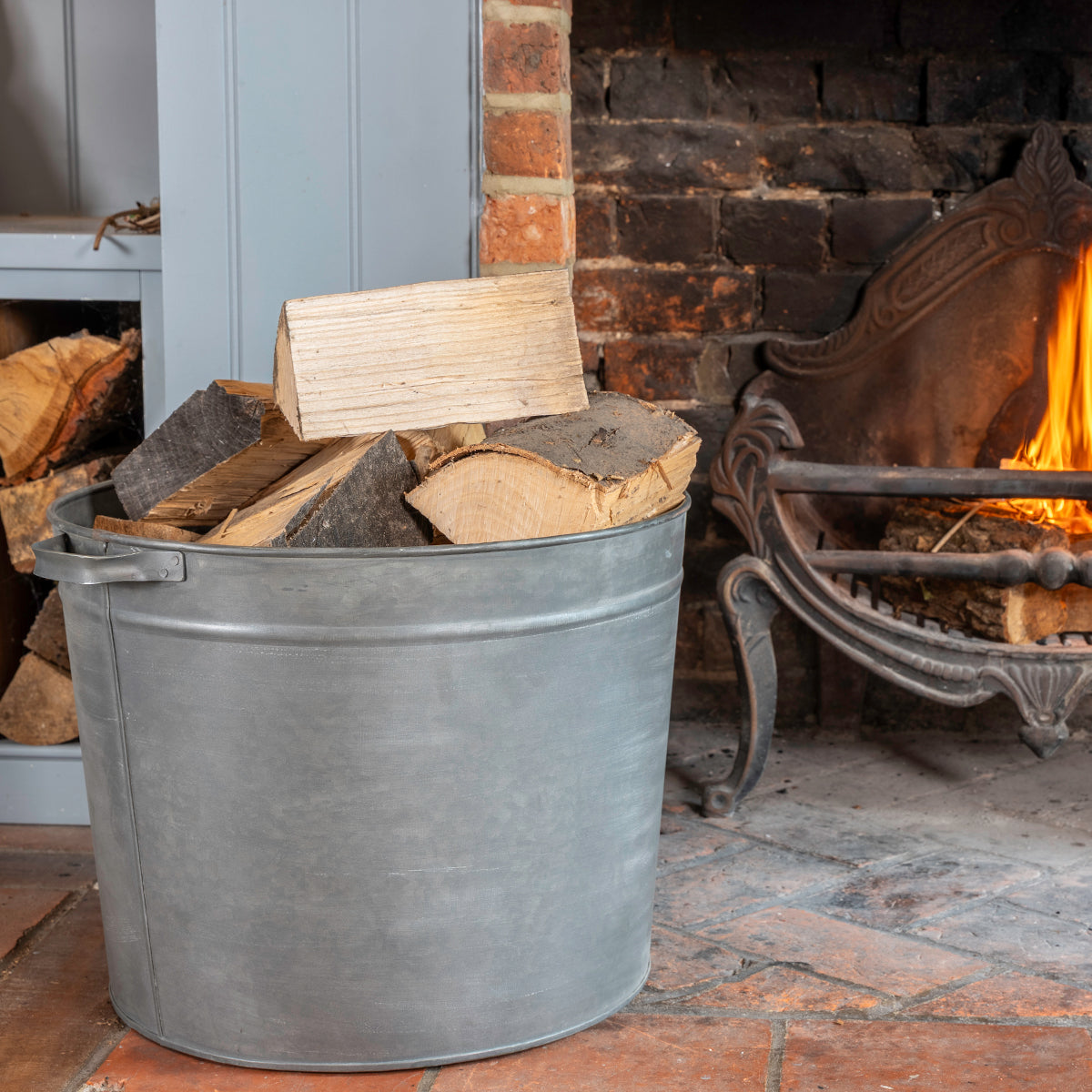 A galvanized steel firewood bucket filled with cut logs, placed in front of a brick fireplace with a burning fire and a wood rack.