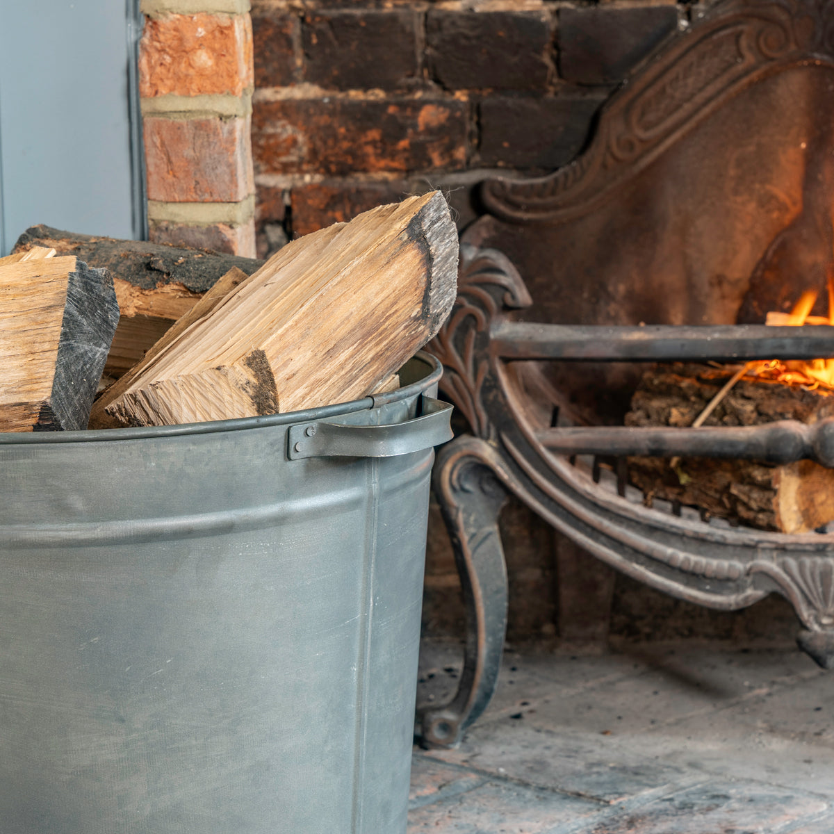 A large gray metal bucket filled with cut firewood logs placed beside a brick fireplace with a burning fire and an ornate cast iron hearth.