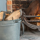 A large gray metal bucket filled with cut firewood logs placed beside a brick fireplace with a burning fire and an ornate cast iron hearth.