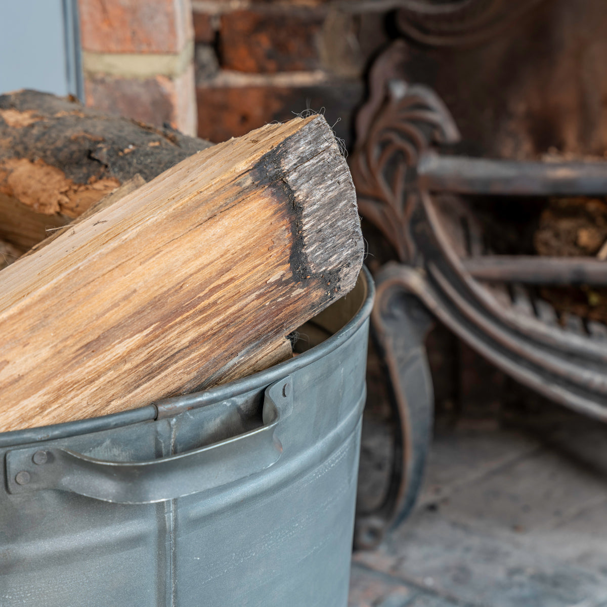A gray metal bucket filled with cut firewood logs, placed in front of a rustic fireplace with a metal grate and brick hearth.